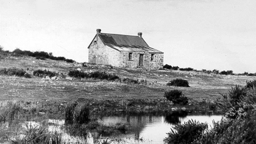 Black and white photo of an old stone house on a hill with a small pond in front of it