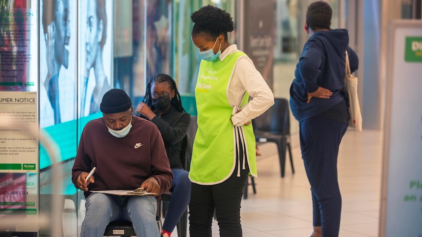 A female nurse in protective clothing stands over a man sitting writing on a clipboard