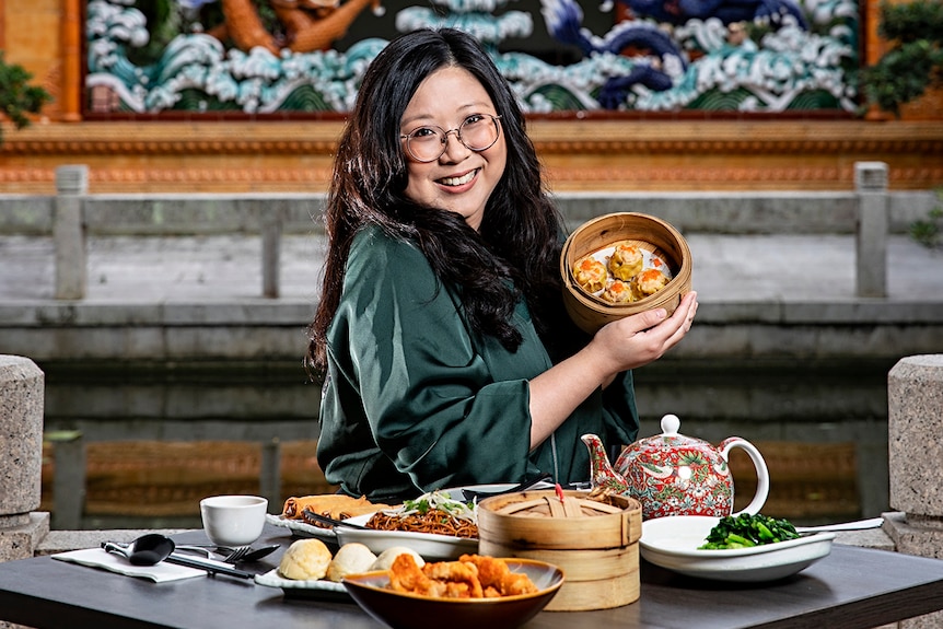 A Chinese woman in her 30s sittings in a Chinese restaurant holding up a steam basket of dumplings
