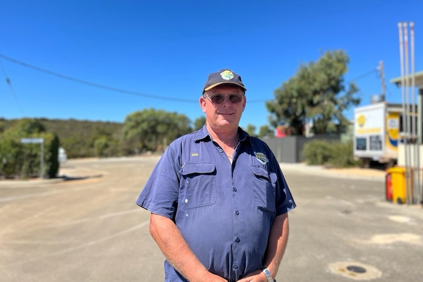 An older man in blue stands on a road in sunshine