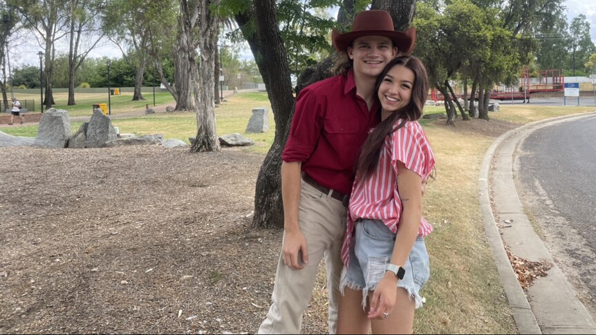Tyla in a pink and white shirt, Jarrad in a hat and red shirt leaning into each other smiling in front of a tree