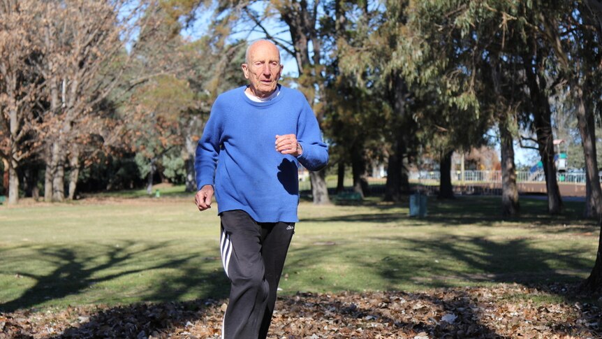 Elderly man runs in park through fallen autumn leaves.