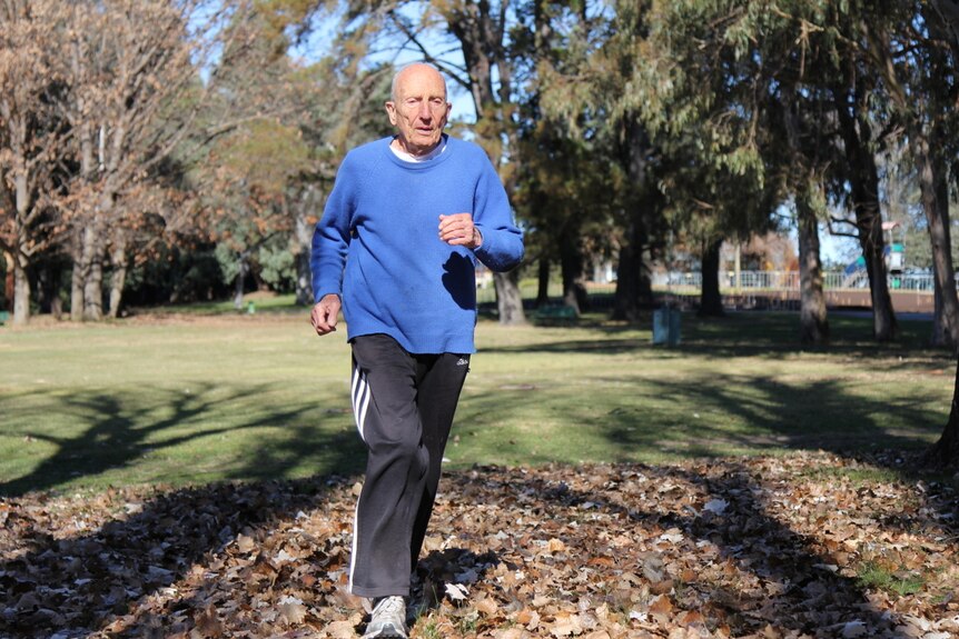 Elderly man runs in park through fallen autumn leaves.