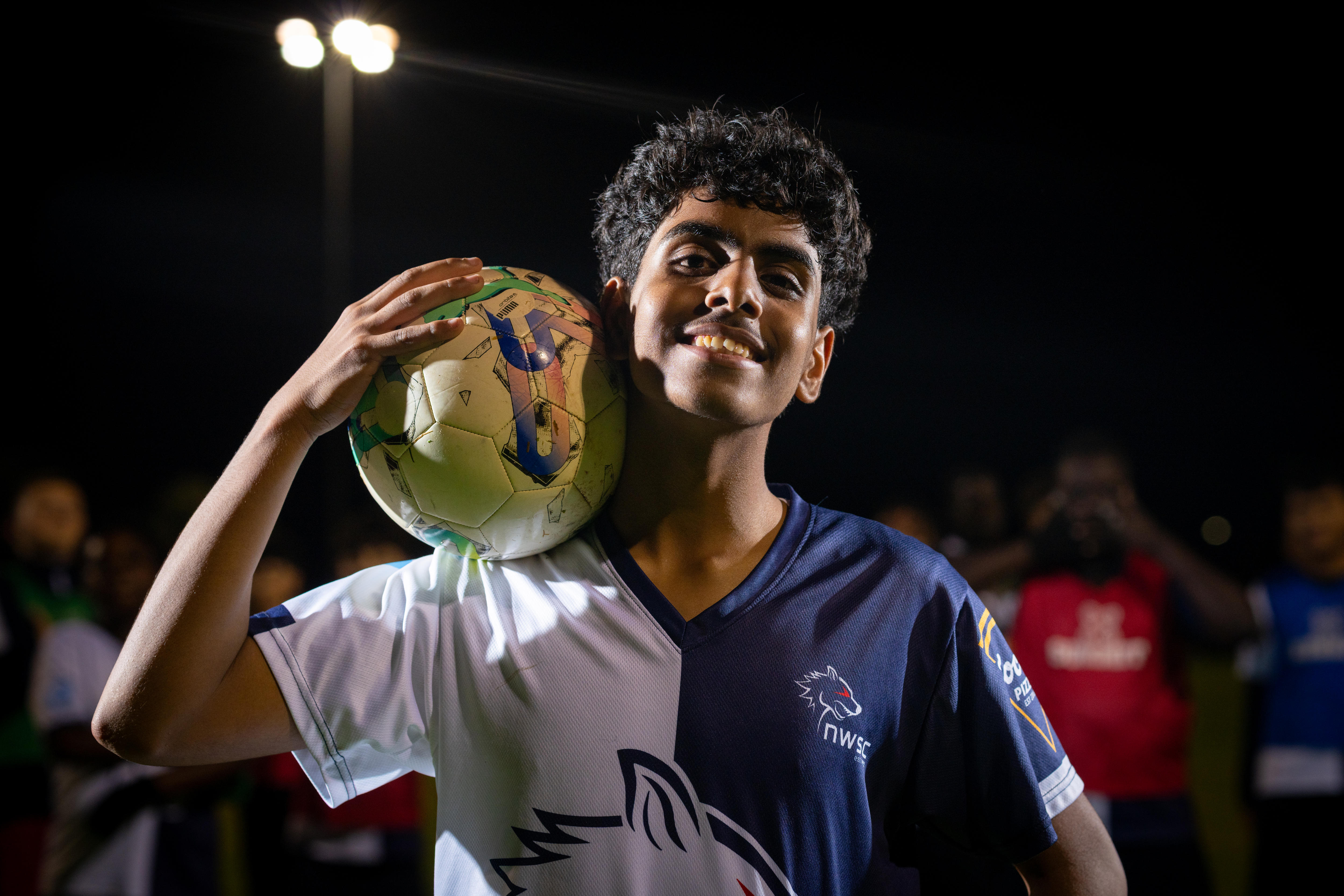 A boy in a soccer uniform holds a soccer ball on his shoulder.
