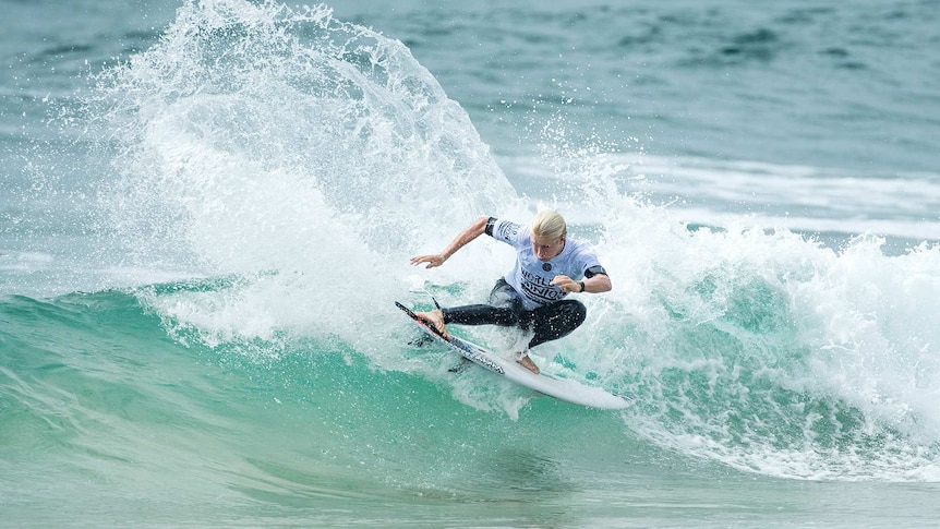 Ethan Ewing crouches low as he surfs a wave, causing a big splash behind him. 