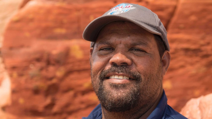 Close-cropped portrait of an indigenous man wearing a baseball cap, standing in front of some rocks.