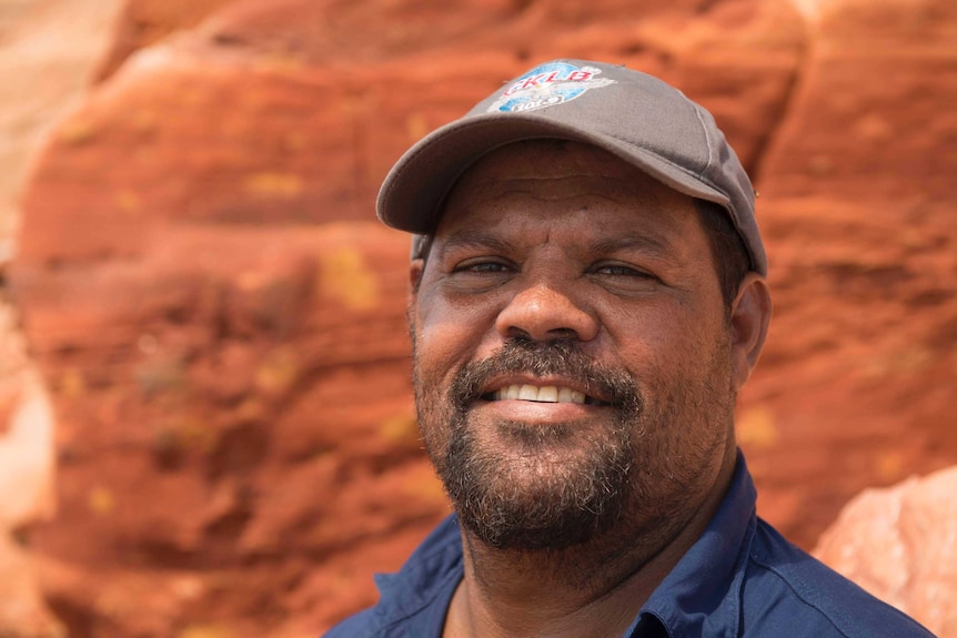 Close-cropped portrait of an indigenous man wearing a baseball cap, standing in front of some rocks.