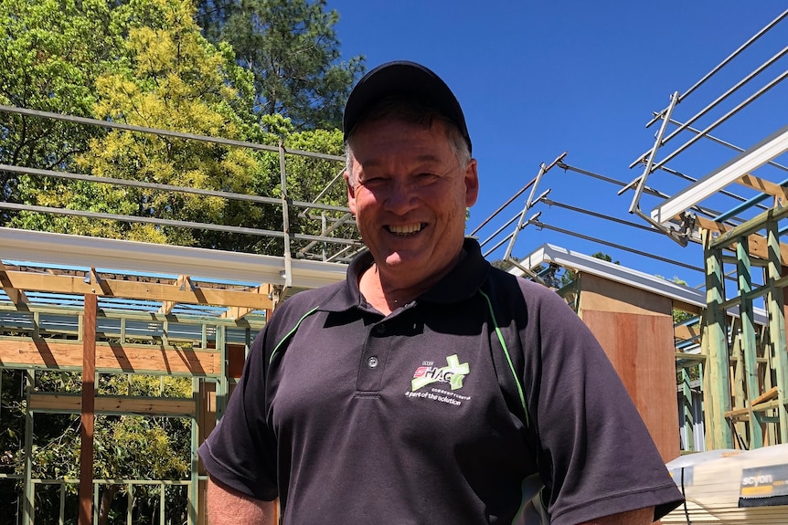Man with cap, polo shirt and jeans smiles into sun as tiny house frames are built behind him