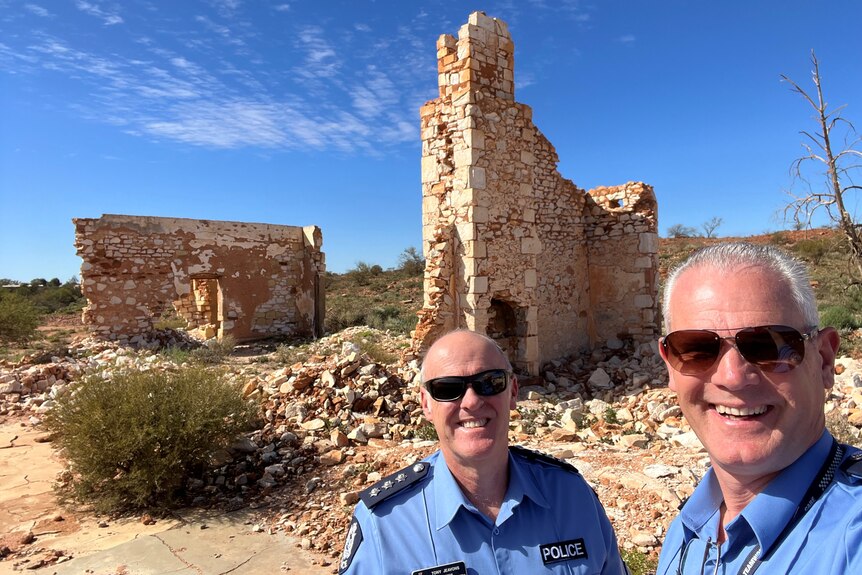 Two smiling middle aged white, balding men in police uniform outside ruins under blue skies. 