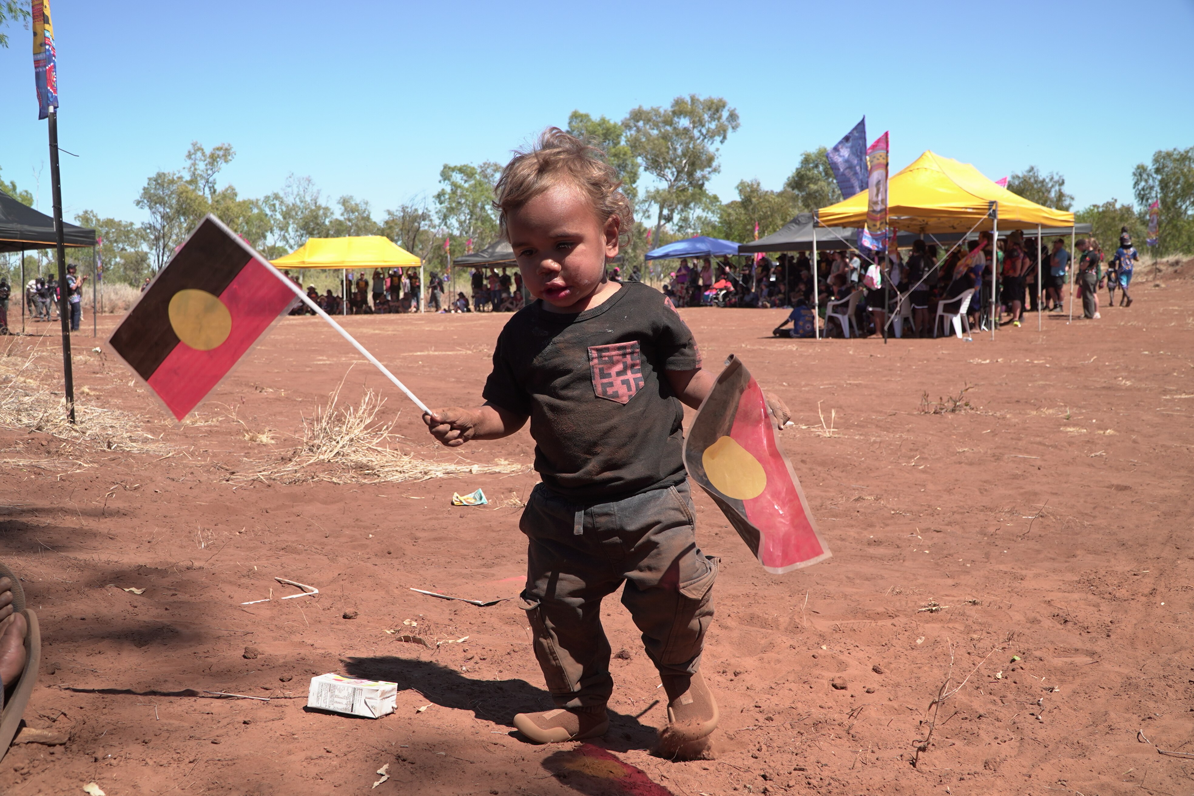 Small Aboriginal toddler dancing on dirt ground, wearing black shirt and pants, holding two small Aboriginal flags.
