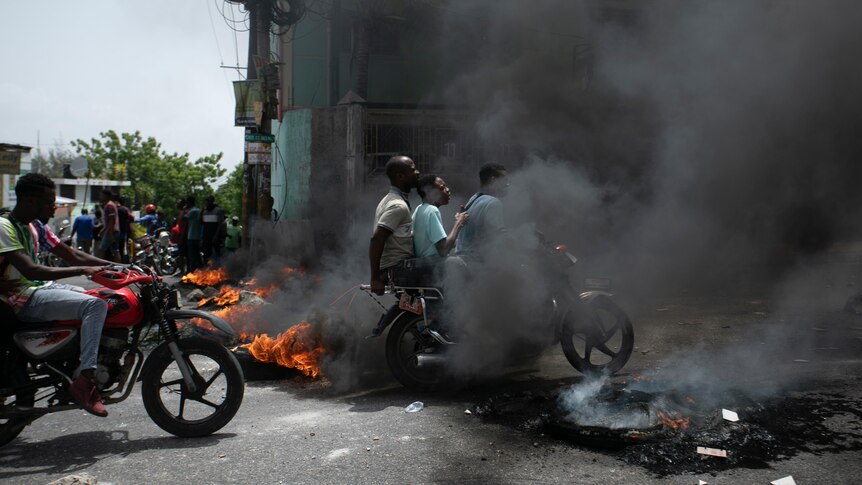 A motorcycle taxi driver carries clients past a burning barricade.