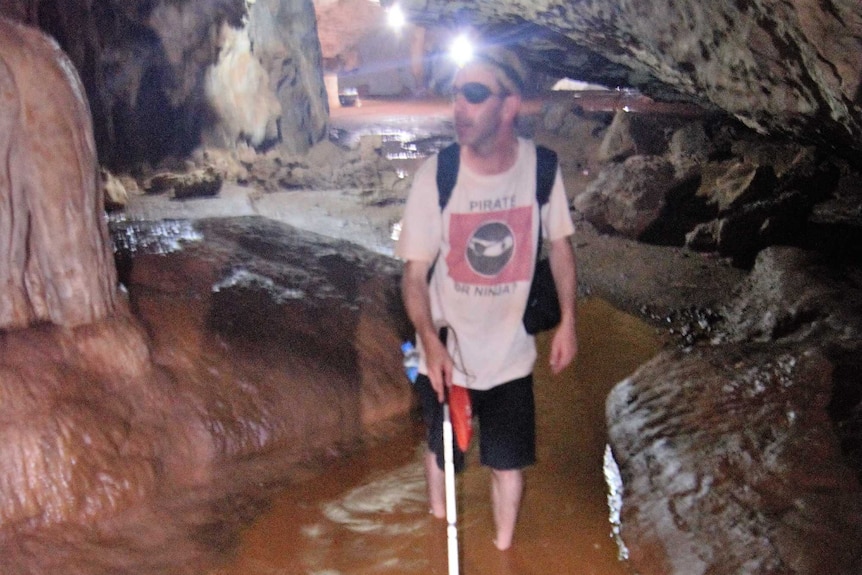 A man walks in shin-deep water, in a dark cave in Myanmar using a white cane for guidance