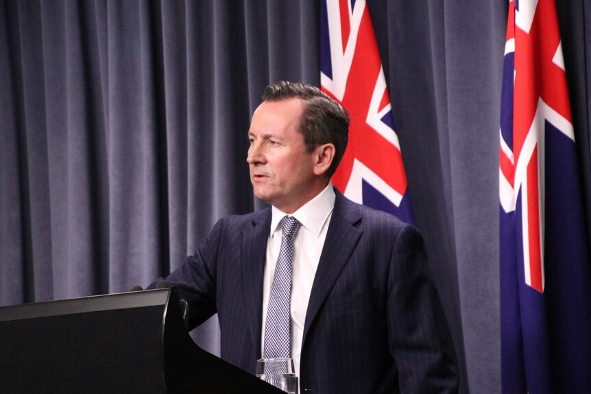 Mark McGowan wearing a blue suit and blue and white checked tie, standing at a lectern in front of blue curtains and flags.