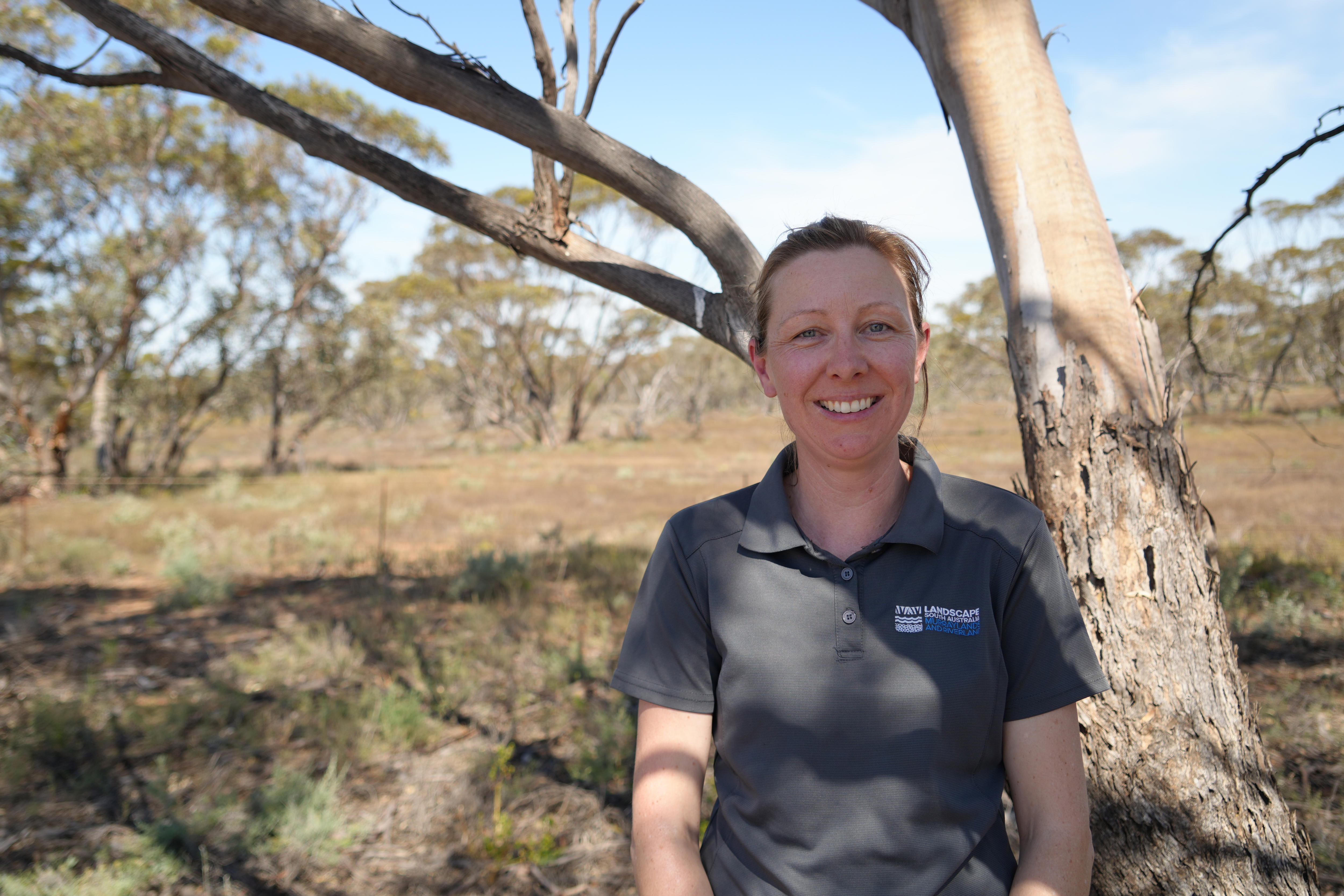A woman wearing a polo shirt stands in front of gum tree and native Australian vegetation.