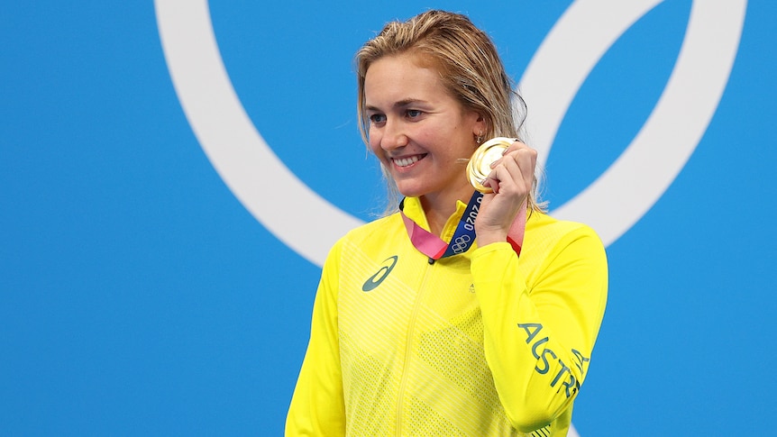 A blonde woman wearing a yellow jacket stands holding a gold medal