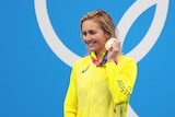 A blonde woman wearing a yellow jacket stands holding a gold medal