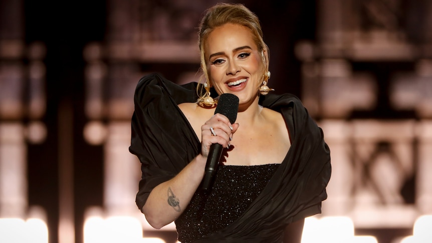 Adele singing into a microphone, with a Saturn tattoo on her forearm and Saturn earrings. 