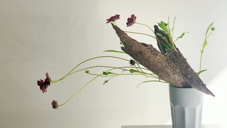 A vase with an arrangement of flowers and bark is on a pedestal with a white background.