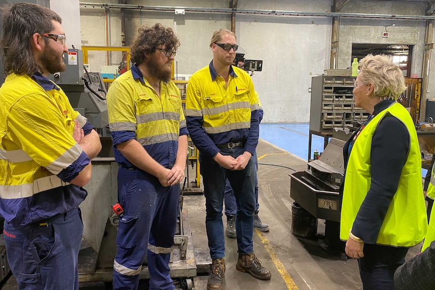 Three young men in high vis work wear talk to a female politician while standing in a workshop.