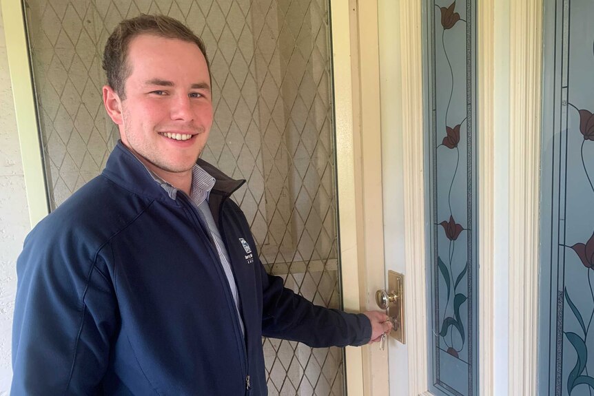 A young man in a navy jacket holds a key in a wooden door, smiling.