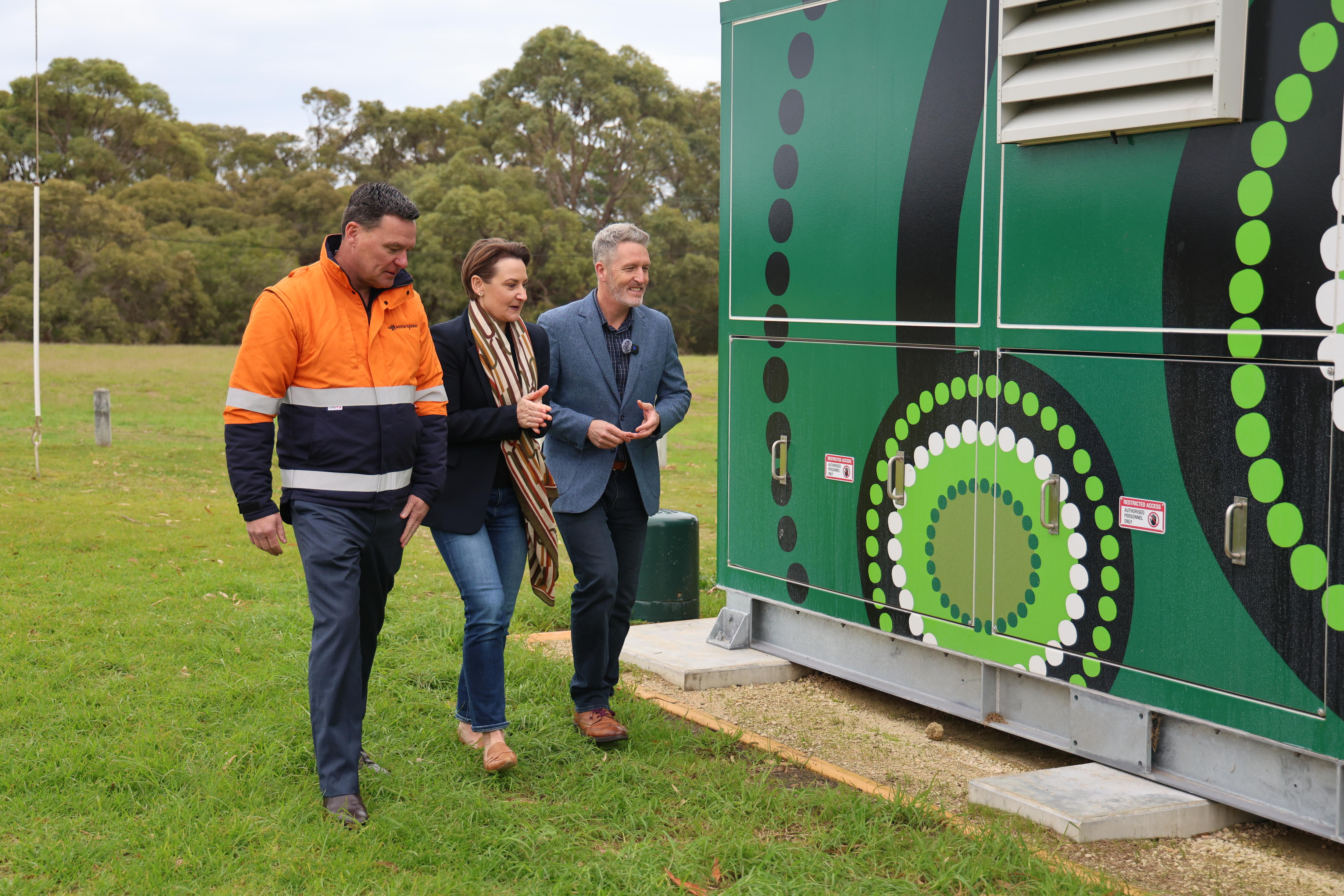 Three people walk past a community battery painted with green artwork.