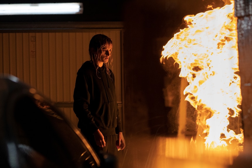 A young woman stands in front of a wall of flames