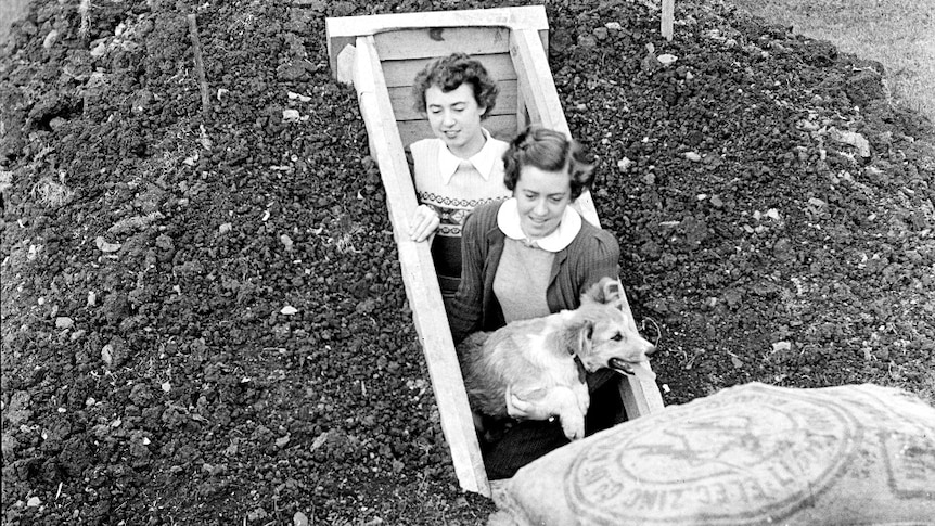 A black and white image of two women in 1940s clothes coming out of a hole in the ground, one holding a pet dog