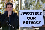 Senator Ludlam stands next to a man in an 'Anonymous' mask, holding a sign saying '#protect our privacy'.