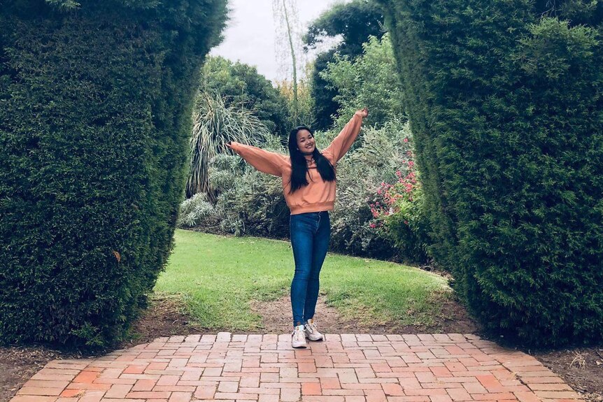 A picture of a woman in jeans and a sweater with her arms in the air between two giant green hedges.
