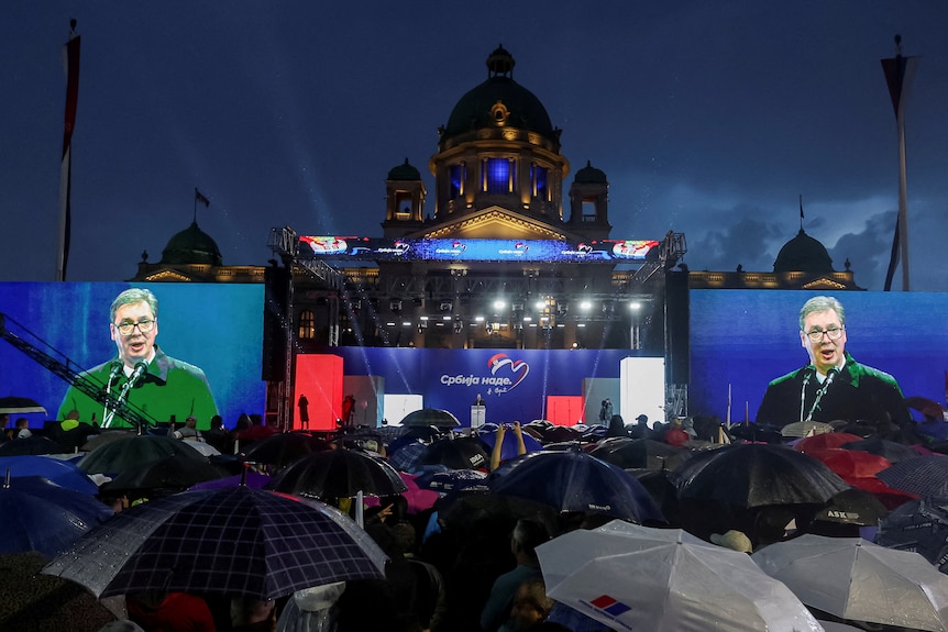 People with umbrellas stand in front of a stage and two large video screens showing a white, middle-aged man giving a speech.