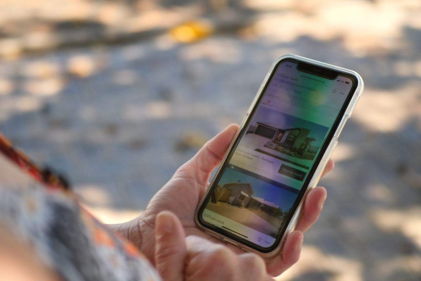 A woman holds a mobile phone, on the screen you can see pictures of houses on a rental housing website.