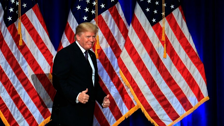 Donald Trump gives two thumbs up in front of American flags.