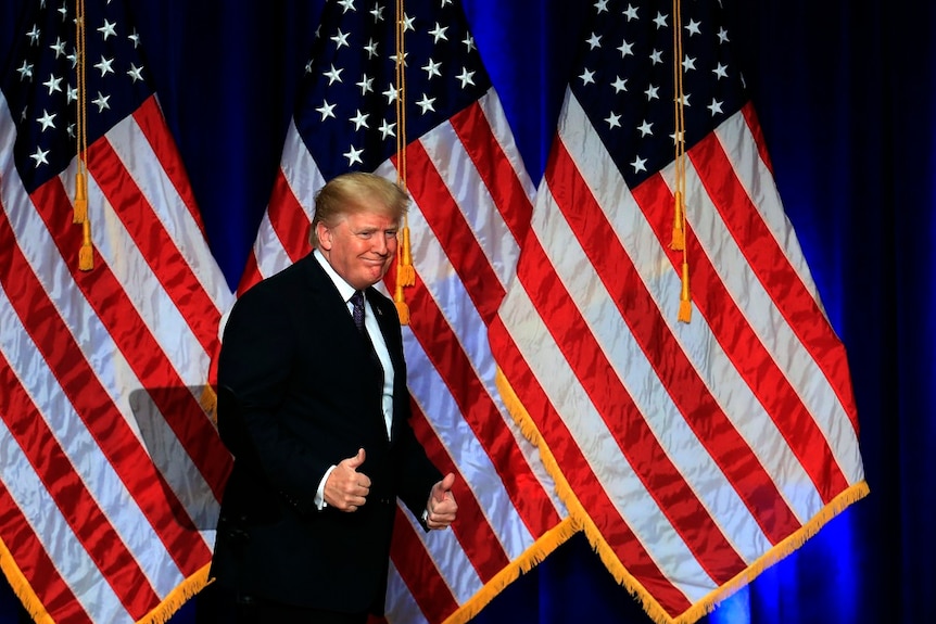 Donald Trump gives two thumbs up in front of American flags.