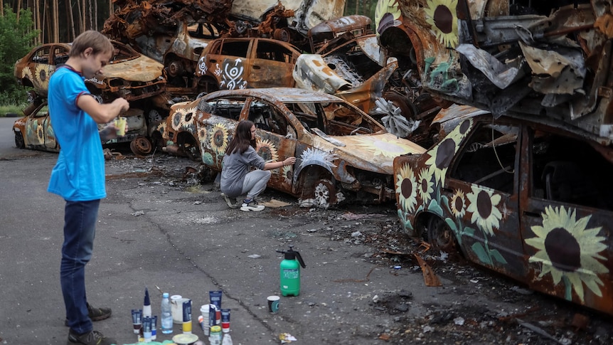 On the right, a pile of destroyed cars is pictured with sunflowers painted on it. On the left an artist looks at his paintwork