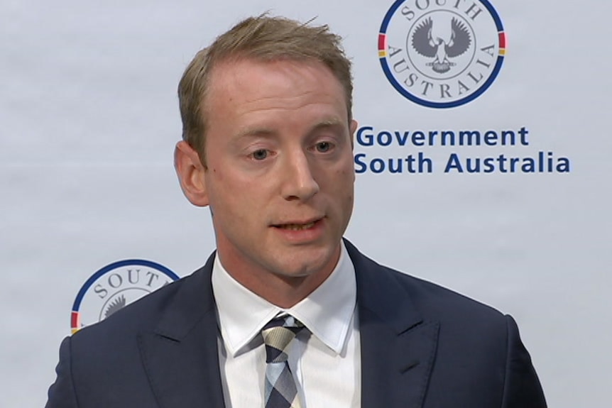 A man wearing a suit and tie speaks in front of an SA government banner