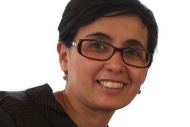 A woman with short hair wearing a dark frame glasses, black blouse and earrings.