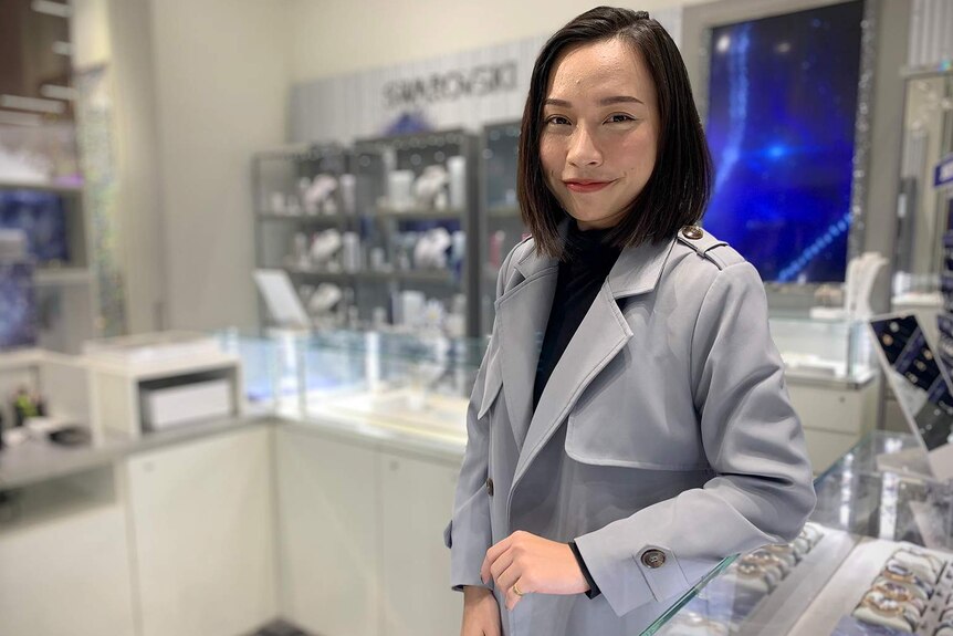 A smiling Vietnamese woman wearing a coat jacket stands in a jewellery store with her arm resting on the counter.