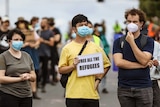 A crowd of people, many wearing masks and standing apart from eachother, with one holding a sign saying 'free all the refugees'.