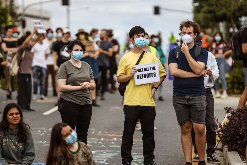 A crowd of people, many wearing masks and standing apart from eachother, with one holding a sign saying 'free all the refugees'.