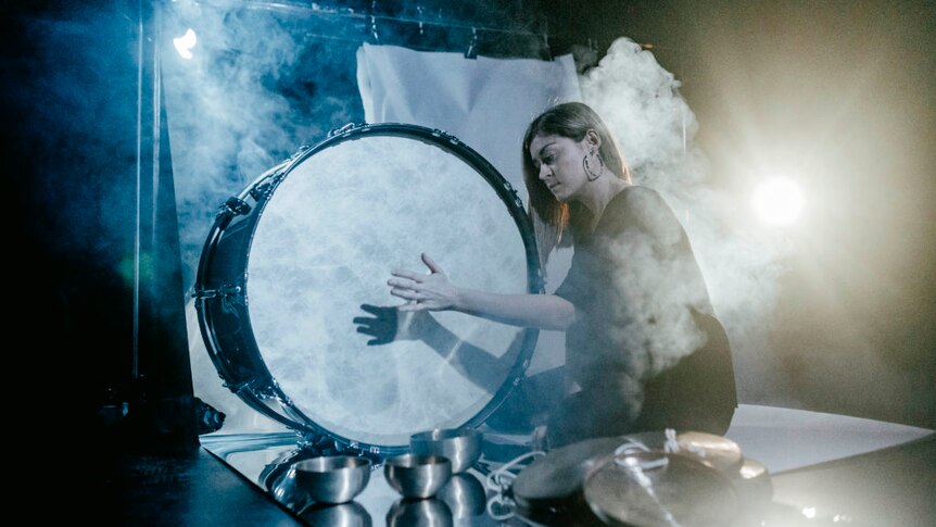 Louise sits on a stage surrounded by smoke hitting a large drum with her hand.