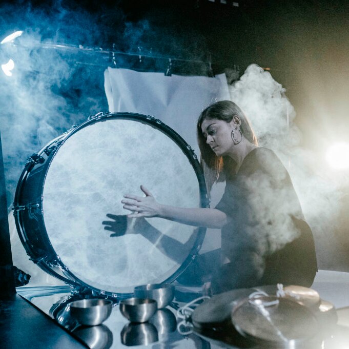 Louise sits on a stage surrounded by smoke hitting a large drum with her hand.