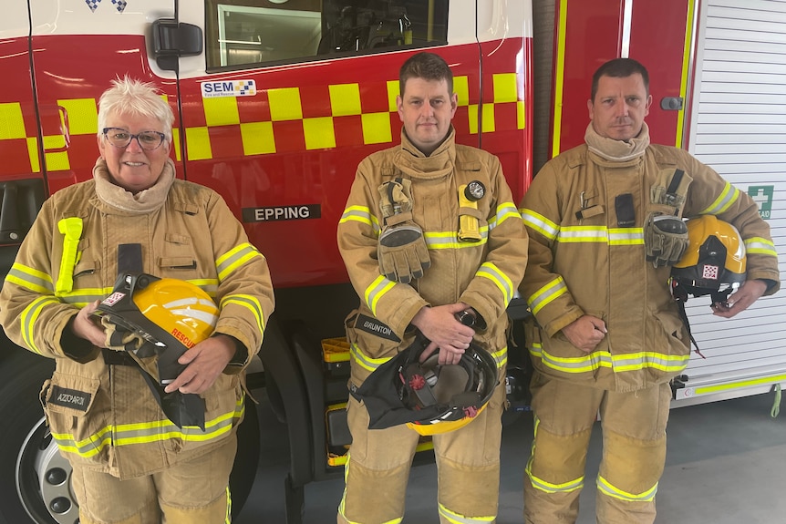 Three firefighters stand in front of a firetruck.