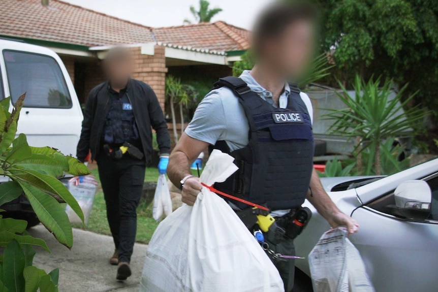 Police officers with their faces blurred hold large white sacks outside a house.