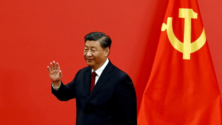 Xi Jinping gives a little wave next to a Chinese flag