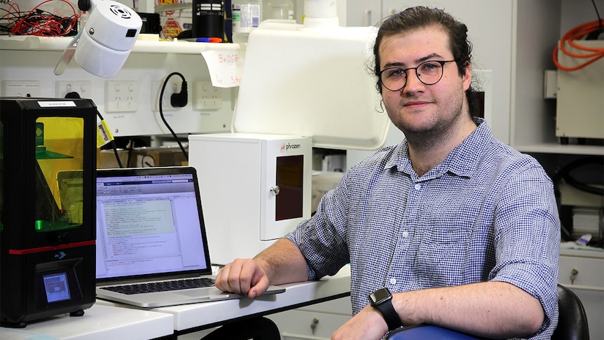 A young, bespectacled man with long, dark hair sits in a university lab.