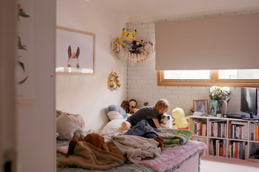 A wide shot of a young woman sitting on her bed with her dog. Her room is full of stuffed toys, books, and blankets. 