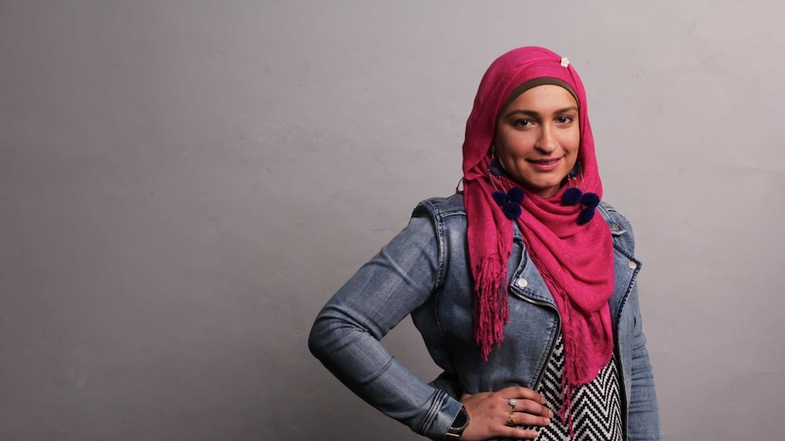 A young woman wearing a bright pink head scarf and acid-wash denim jacket smiles, posing against a grey backdrop.