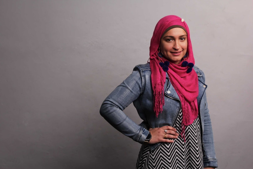 A young woman wearing a bright pink head scarf and acid-wash denim jacket smiles, posing against a grey backdrop.