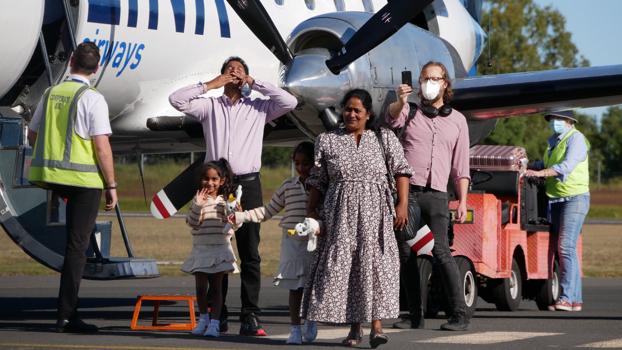 A family stands on tarmac beside a plane and emotionally greets a crowd.