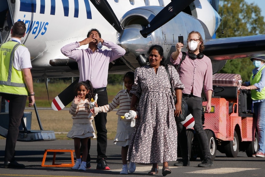 A family stands on tarmac beside a plane and emotionally greets a crowd.